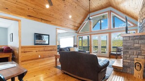 A spacious living room with wooden floors, a stone fireplace, and large windows offering a view of the outdoors.