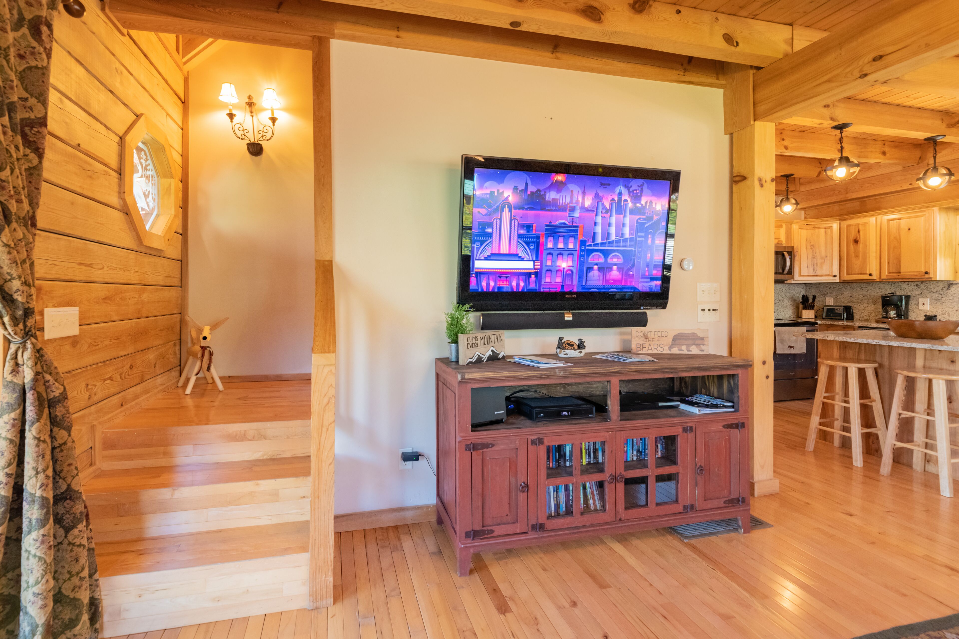 A wooden interior with a TV on a wooden stand, a kitchen area, and a staircase.
