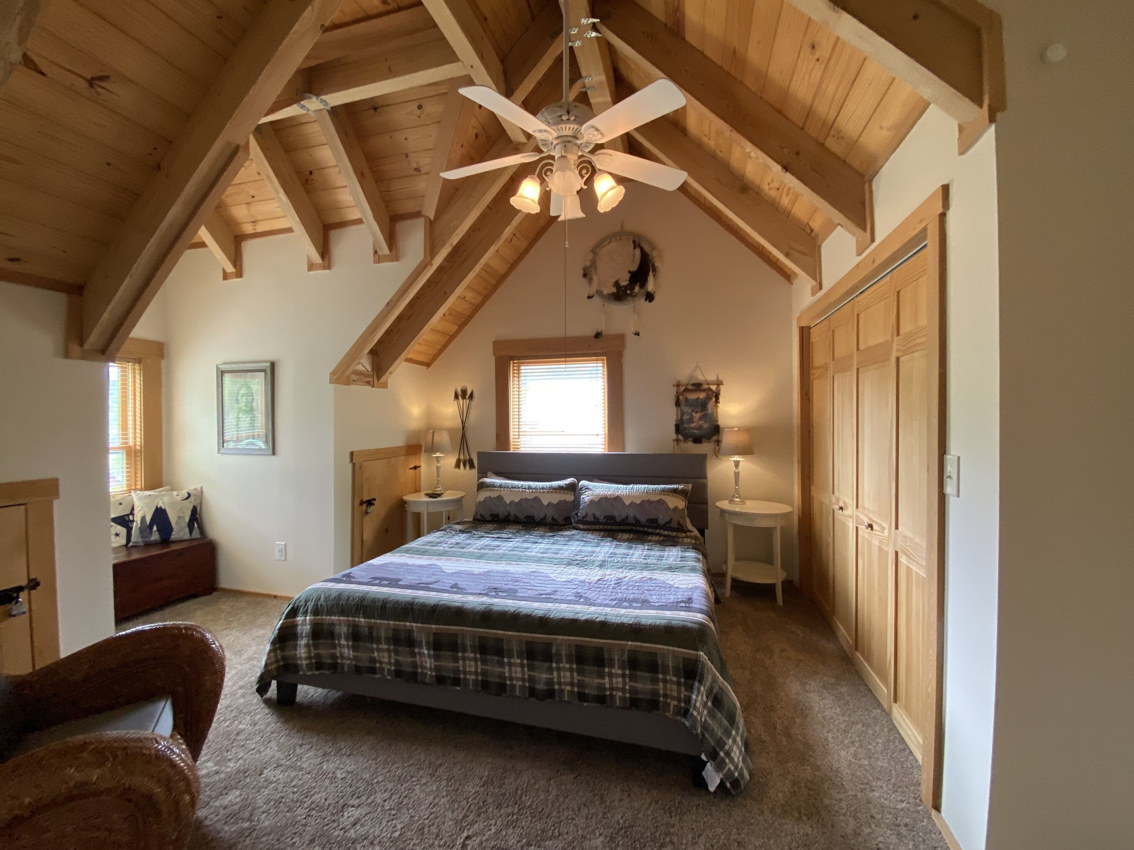 A bedroom with a large bed, wooden ceiling beams, a ceiling fan, and a window with blinds.