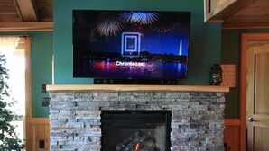 A fireplace with a stone surround and a flat-screen TV mounted above it displaying fireworks. - ~ Lakeside Log Cabin ~ Blessings & Memories Abound! (Graham)
