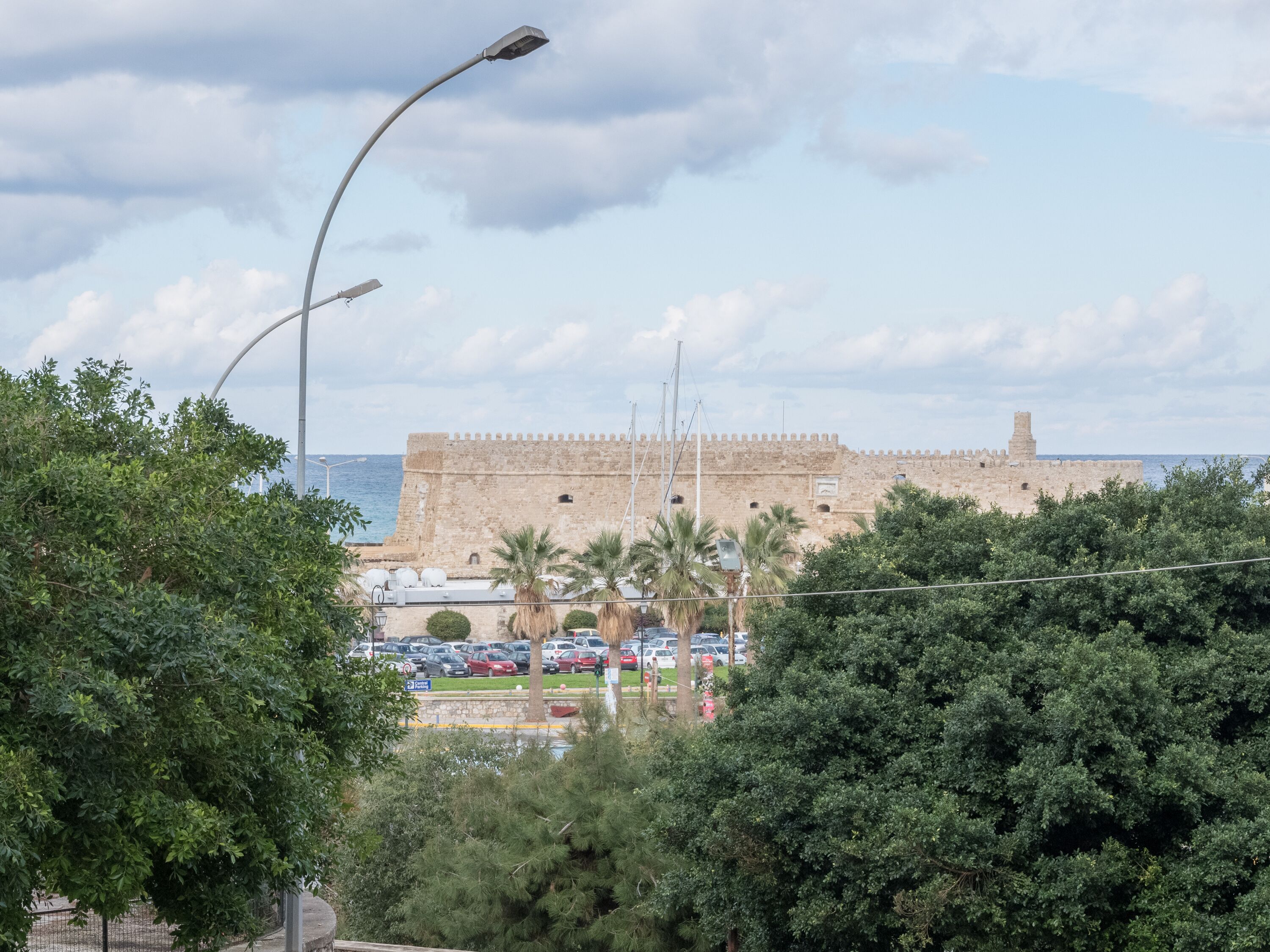 Un edificio de piedra histórico con una muralla fortificada, coches aparcados y un farol en primer plano.