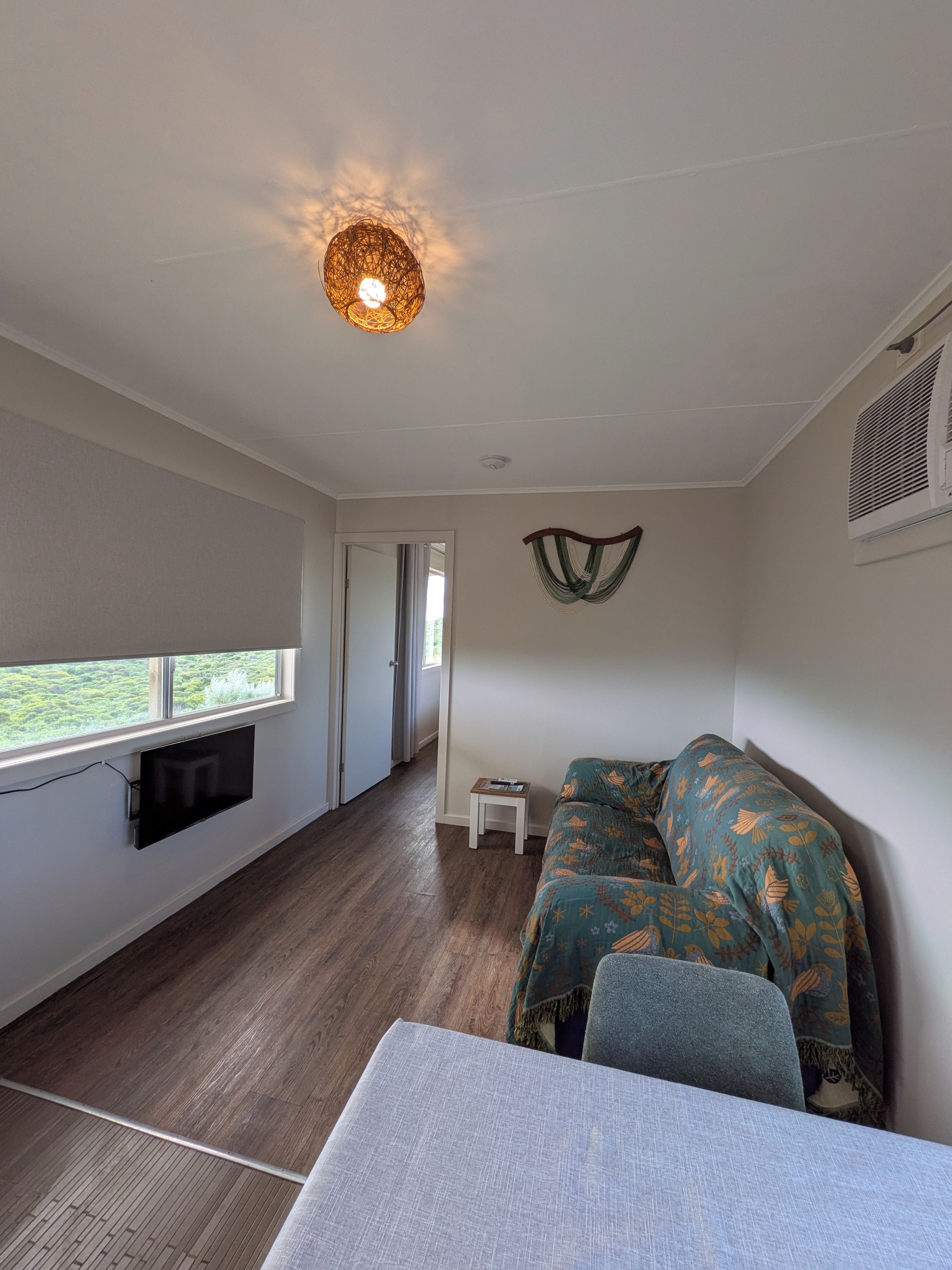 Een kamer met een wandgemonteerde flatscreen-tv, een tafeltje, een bank met gestreepte kussens, een plafondlamp en een raam dat voor natuurlijk licht zorgt.