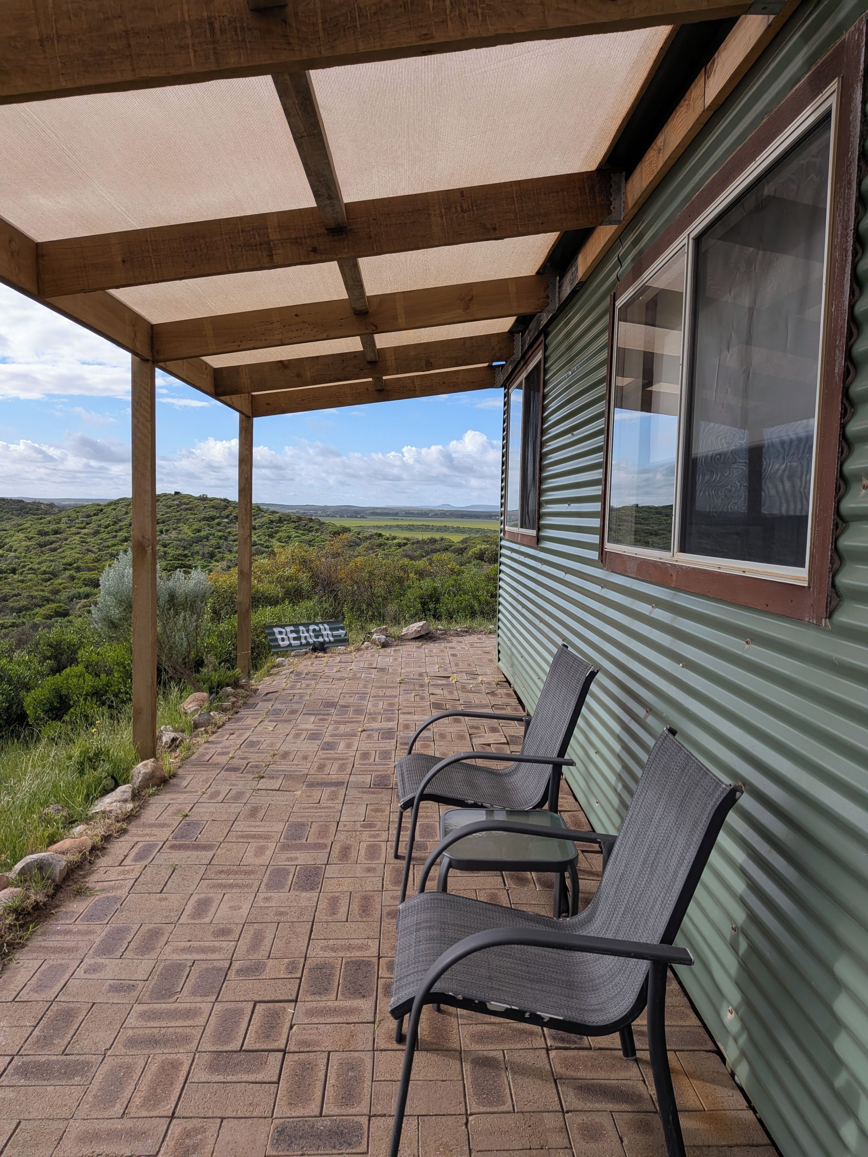 Ett takförsett utomhusområde med sittplatser, bestående av två stolar och ett bord, en skylt med texten ”STRAND” och utsikt över ett grönskande landskap.