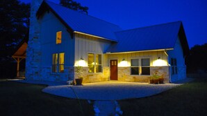 A stone house with a steep gabled roof, a front porch with potted plants, and a lit entrance. - Private Riverfront Lodge with Mountain Views Between Lost Maples and Garner Park (Vanderpool)