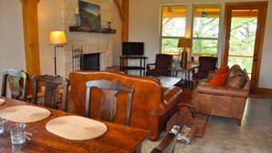 A cozy living room with a fireplace, wooden furniture, and large windows offering a view of the outdoors. - Private Riverfront Lodge with Mountain Views Between Lost Maples and Garner Park (Vanderpool)