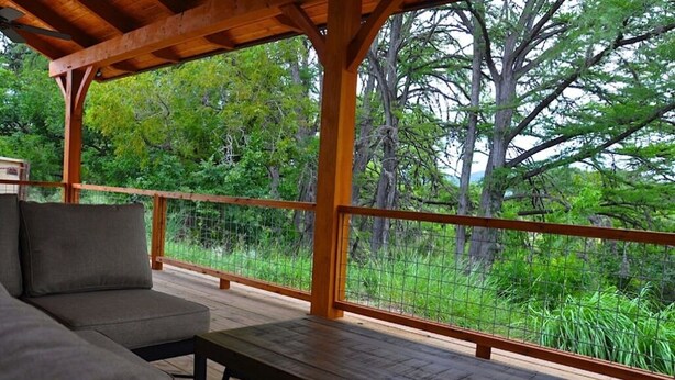 A wooden deck with a sofa and table, surrounded by a metal railing, overlooking a dense forest. - Private Riverfront Lodge with Mountain Views Between Lost Maples and Garner Park (Vanderpool)