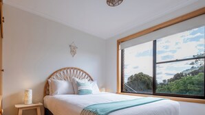Ein Schlafzimmer mit großem Fenster, einem Bett mit weißer Bettwäsche, einem Holzkopfende und einem kleinen Nachttisch mit Lampe.