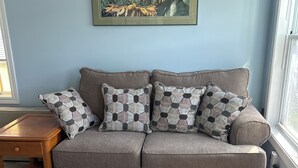 A living room with a grey sofa, patterned cushions, a wooden side table, and a framed artwork on the wall. - Carry Bay Cozy Cottage-Lake Champlain Cabin 4 (North Hero)