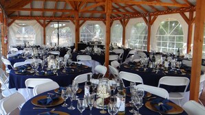 Un espace de restauration aménagé sous une tente, avec des tables rondes dressées pour un événement formel, des chaises blanches et des nappes bleues.