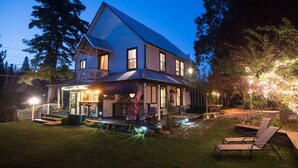Une maison à deux étages avec un porche, un espace salon extérieur et un jardin bien éclairé au crépuscule.