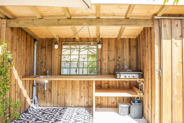 A compact wooden cabin with a small kitchen area, including a sink, a gas stove, and a window with a view of greenery outside. - Maison avec terrasse à 2 minutes de la plage (Arcachon)