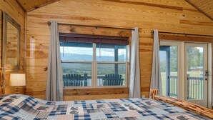 Een houten kamer met een bed, een raam met uitzicht op stoelen buiten, en een deur aan de rechterkant.