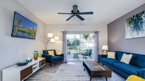 Een moderne woonkamer met een blauwe bank, een gele fauteuil en een salontafel. Er is een tv aan de muur bevestigd en een plafondventilator.