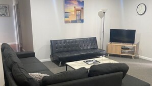 Uma sala de estar com sofá seccional, uma TV de tela plana em suporte de madeira, um quadro na parede e um abajur.