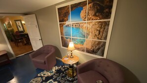 Habitación de hotel con un ventanal amplio que ofrece vistas al mar, dos sillones morados, un tapete estampado y una mesita con una lámpara.