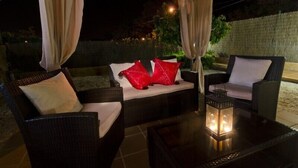 An outdoor seating area with wicker furniture and cushions, a glass table with a lantern, and a covered section with curtains.