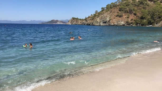 Una playa con agua cristalina, una orilla arenosa y un paisaje montañoso al fondo.