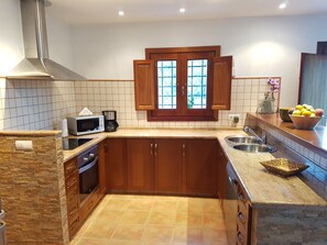 A kitchen with a stone backsplash, wooden cabinets, a stainless steel sink, a microwave, and a window with wooden shutters.