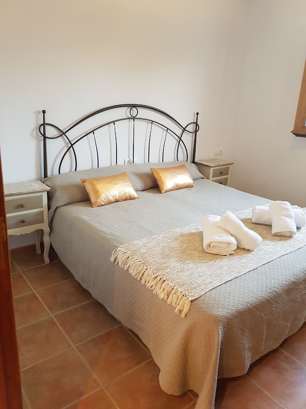 A neatly made bed with a grey bedspread and two gold pillows, a wooden nightstand with a lamp, and a small wooden cabinet.