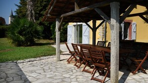An outdoor dining area with a stone-paved patio, wooden tables and chairs, and a yellow building with a stone facade.