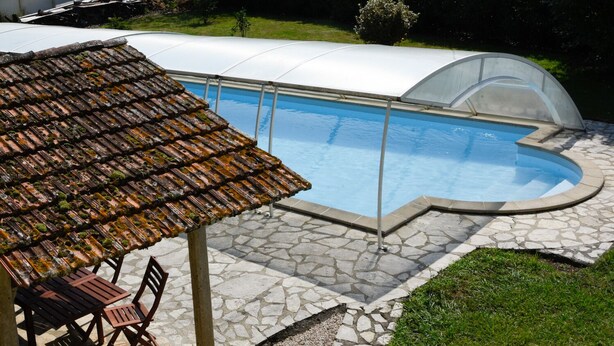 A swimming pool with a stone deck, a covered area with a tiled roof, and a small table with chairs.