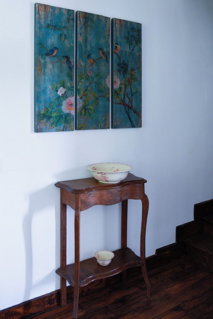 A wooden table with a bowl on it, positioned under a set of three paintings depicting birds and flowers.