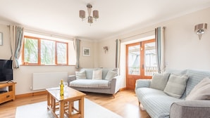A spacious living room with a sofa, a coffee table, and a television. There are windows with curtains, a radiator, and a chandelier. - Duporth Lodge - Three Bedroom Cottage, Sleeps 6 (Charlestown)