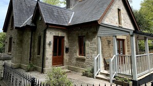 A stone house with a gabled roof, a wooden door, and a porch with a railing. - Duporth Lodge - Three Bedroom Cottage, Sleeps 6 (Charlestown)