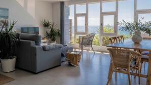 Ein modernes Wohnzimmer mit einem grauen Sofa, einem Esstisch mit Korbstühlen und großen Fenstern mit Meerblick.