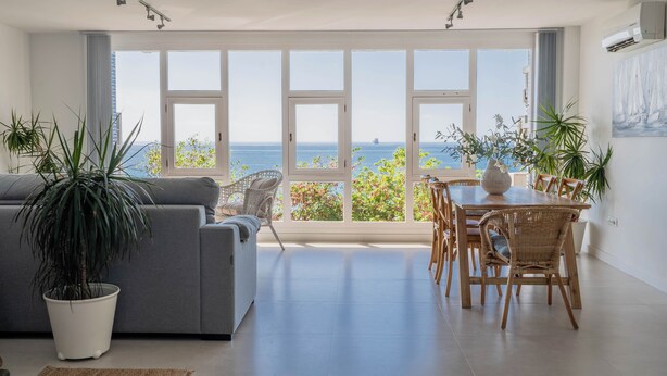 Ein modernes Wohnzimmer mit einem grauen Sofa, einem Esstisch mit Stühlen und großen Fenstern mit Meerblick.