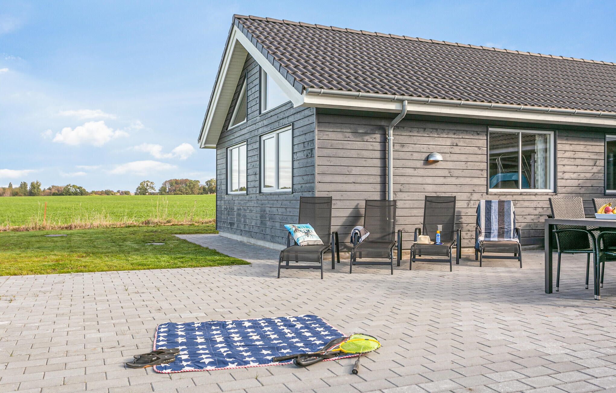 Una casa con techo inclinado, un patio con mobiliario exterior y una cancha de tenis.