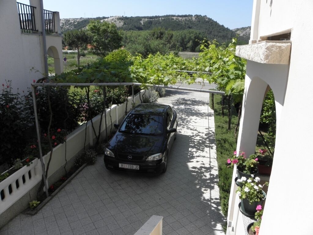 Une voiture noire est garée dans une allée menant à une maison blanche, et un jardin avec des plantes en pot se trouve à proximité.