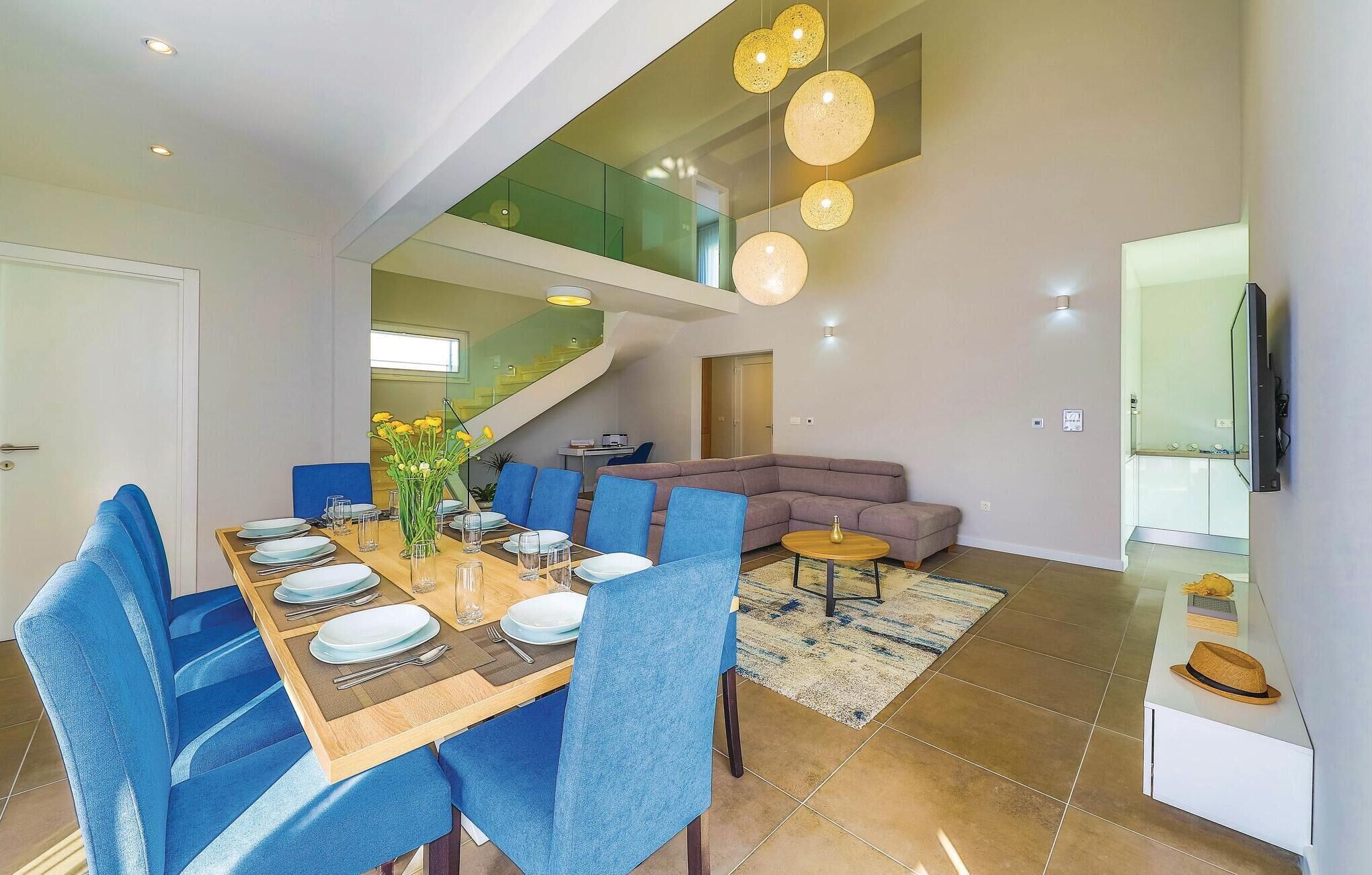 Un espace repas moderne avec une table en bois dressée pour quatre personnes, des chaises à l’assise bleue et un vase rempli de fleurs jaunes. Un escalier mène à un niveau supérieur doté de rampes en verre.