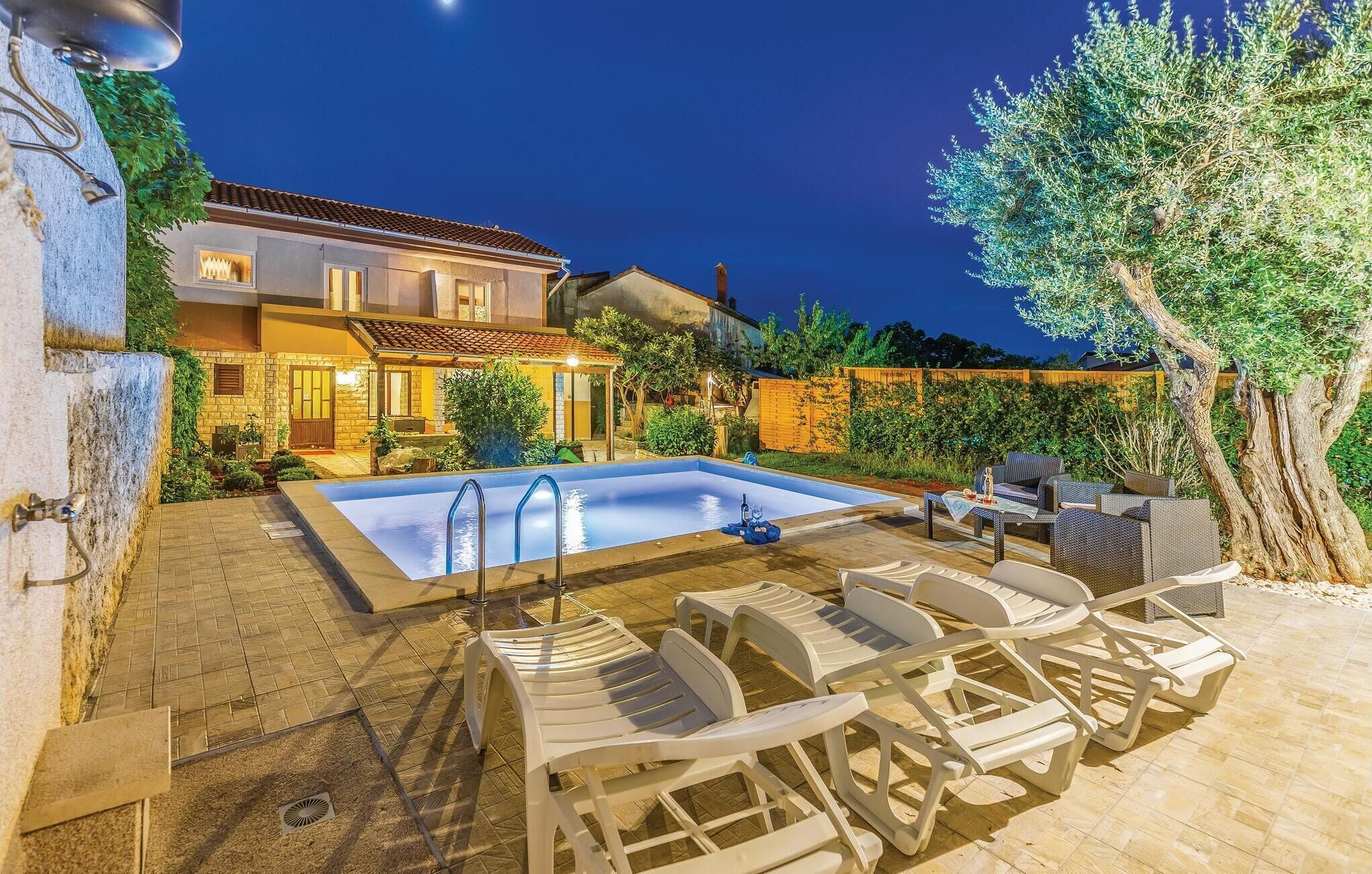 A swimming pool with lounge chairs and a pergola, surrounded by a garden and a house with a tiled roof.
