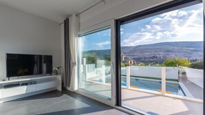 A modern living room with a flat-screen TV, a console table, and a sliding glass door leading to a balcony with a pool and a scenic mountain view. - Awesome home in Srinjine (Srinjine)