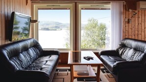 Ein Zimmer mit zwei Ledersofas, die zu einem großen Fenster mit Blick auf einen See und Bäume blicken. Auf einem kleinen Tisch liegen ein Fernseher und ein Notizbuch.