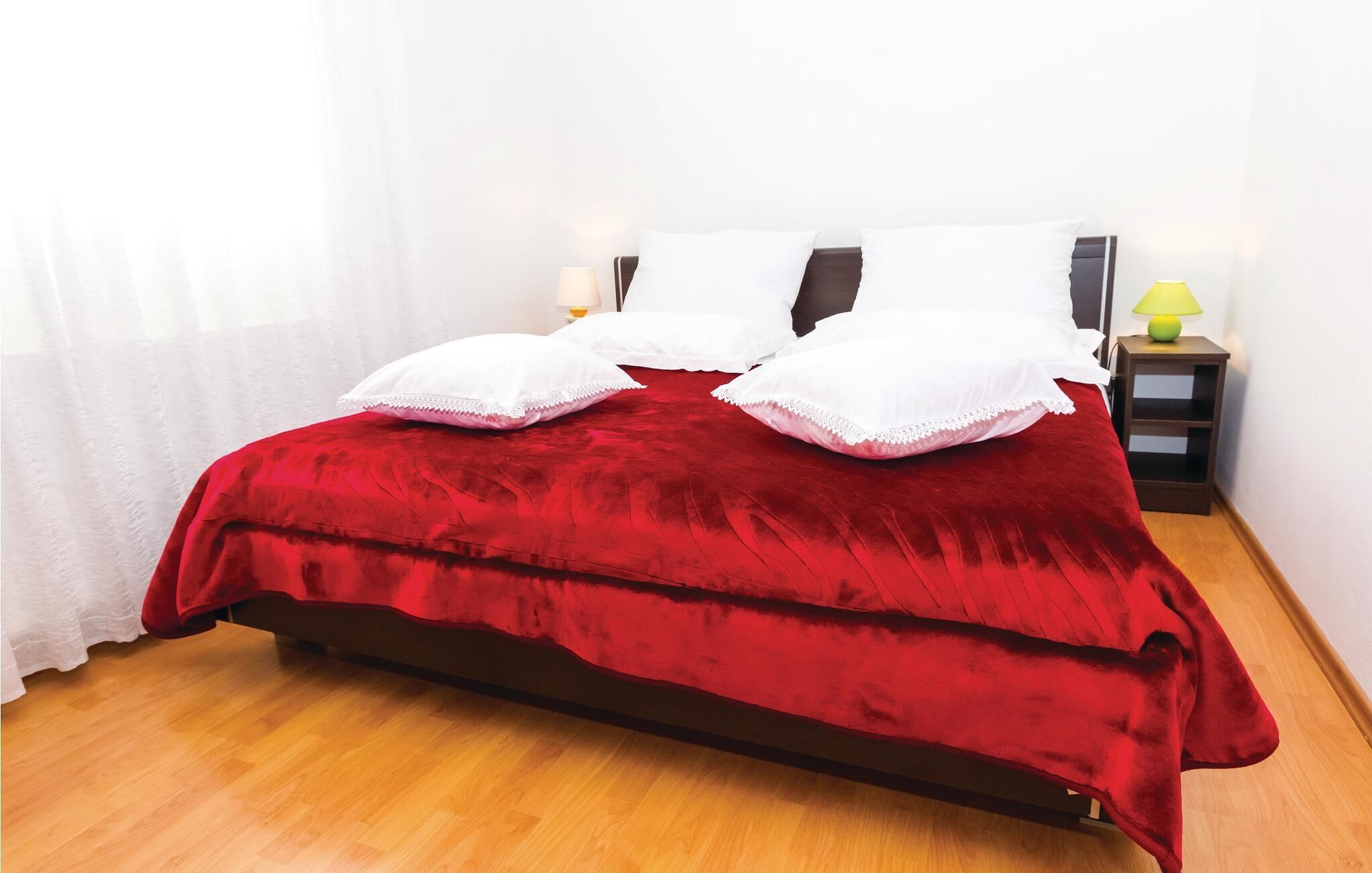 A neatly made bed with a red bedspread and white pillows, a wooden floor, and a nightstand with a lamp.