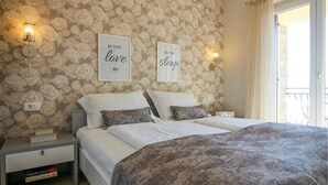 Un dormitorio con papel tapiz floral, dos mesitas de noche con lámparas, una cama con ropa de cama blanca y un edredón estampado, y dos citas motivacionales enmarcadas en la pared.