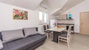 A modern apartment with a grey sofa, a dining table with chairs, a kitchen with a fridge and oven, and a window with a view of the outside. - Lovely apartment in Biograd with WiFi (Biograd)