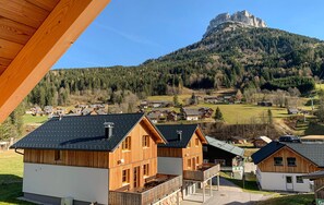 A wooden cabin with a balcony, surrounded by a forested area and a mountain in the background.