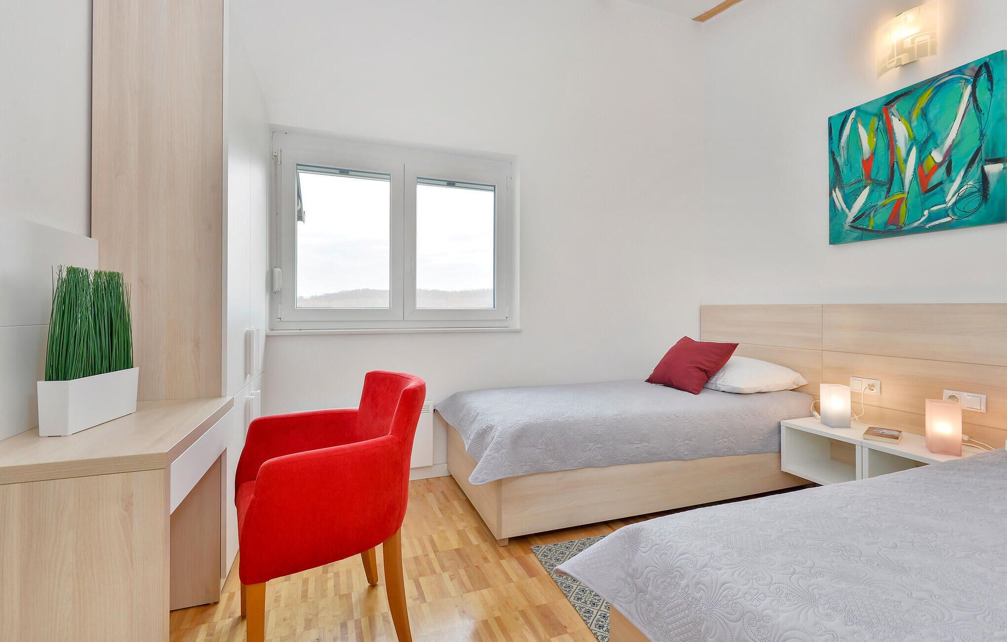 Ett modernt hotellrum med två sängar, ett skrivbord, en röd stol, ett fönster med utsikt och en tavla på väggen.