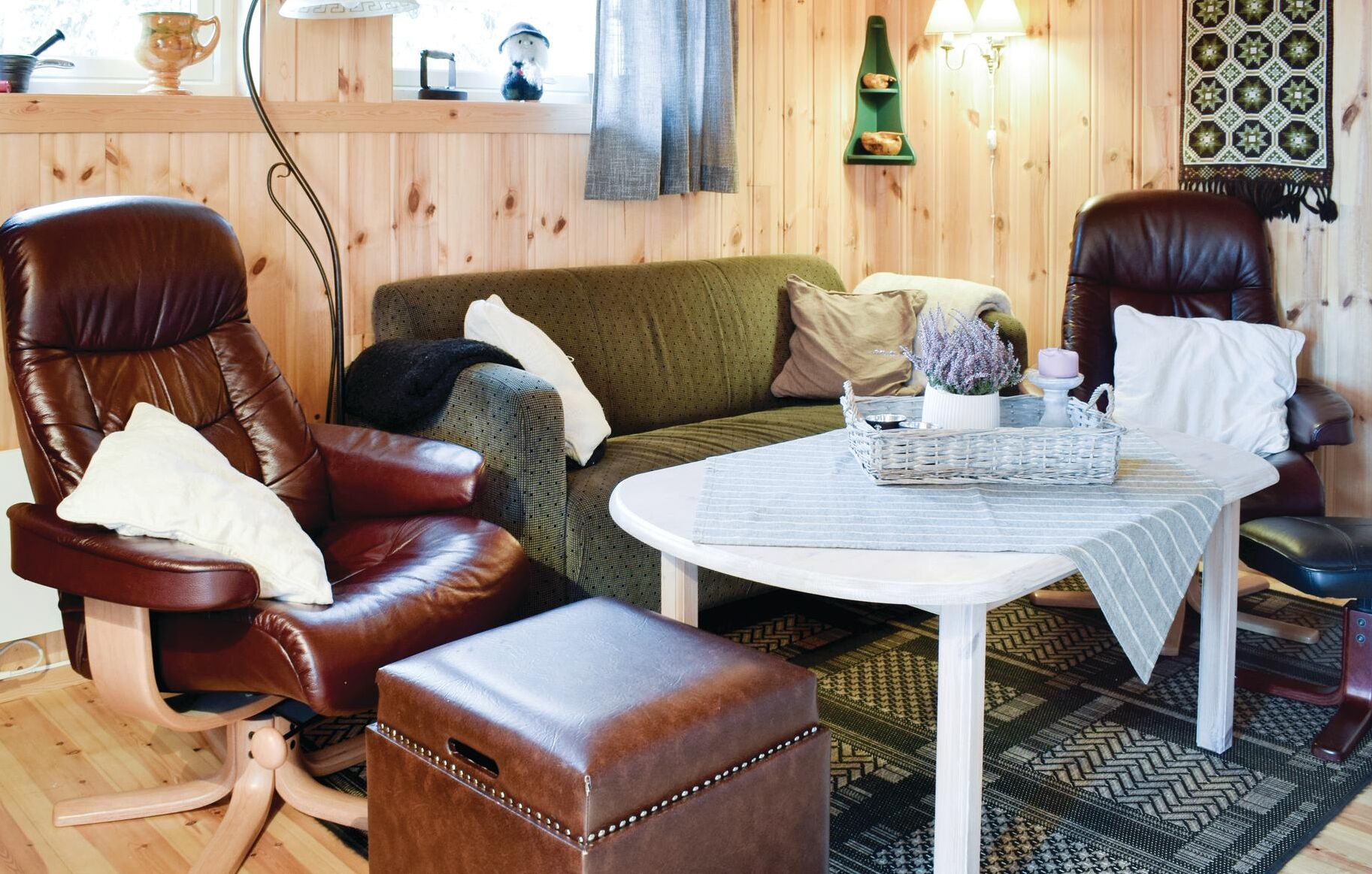 Un salon chaleureux avec un canapé vert, deux fauteuils en cuir, une petite table blanche sur laquelle se trouve un panier, et un tapis à motifs.