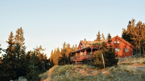 Eine Holzhütte mit Balkon, umgeben von hohen Bäumen und einem grasbewachsenen Hügel.