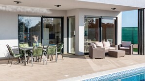 Een modern terras buiten met een zwembad, met een eethoek met stoelen en een tafel, en een zithoek met banken.