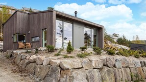 Une cabane moderne en bois, dotée de grandes fenêtres en verre, d’un mur de soutènement en pierre et de mobilier d’extérieur.