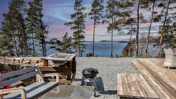Une terrasse en bois équipée d’un barbecue, d’une table et d’un banc, offrant une vue sur un lac bordé d’arbres et sur des montagnes lointaines.