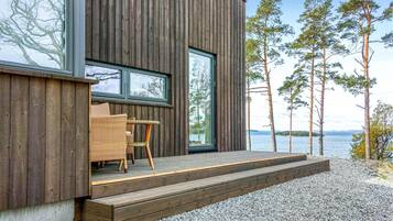 Un bâtiment moderne en bois, doté d’une terrasse en bois, de meubles en osier et offrant une vue sur l’eau et les arbres.