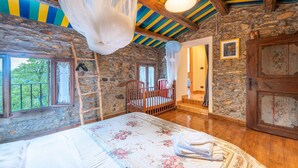 Ein rustikales Schlafzimmer mit Steinwand, Holzbalkendecke, einem Bett mit Blumenmuster-Bettwäsche, einem Babybett und Blick ins Grüne.