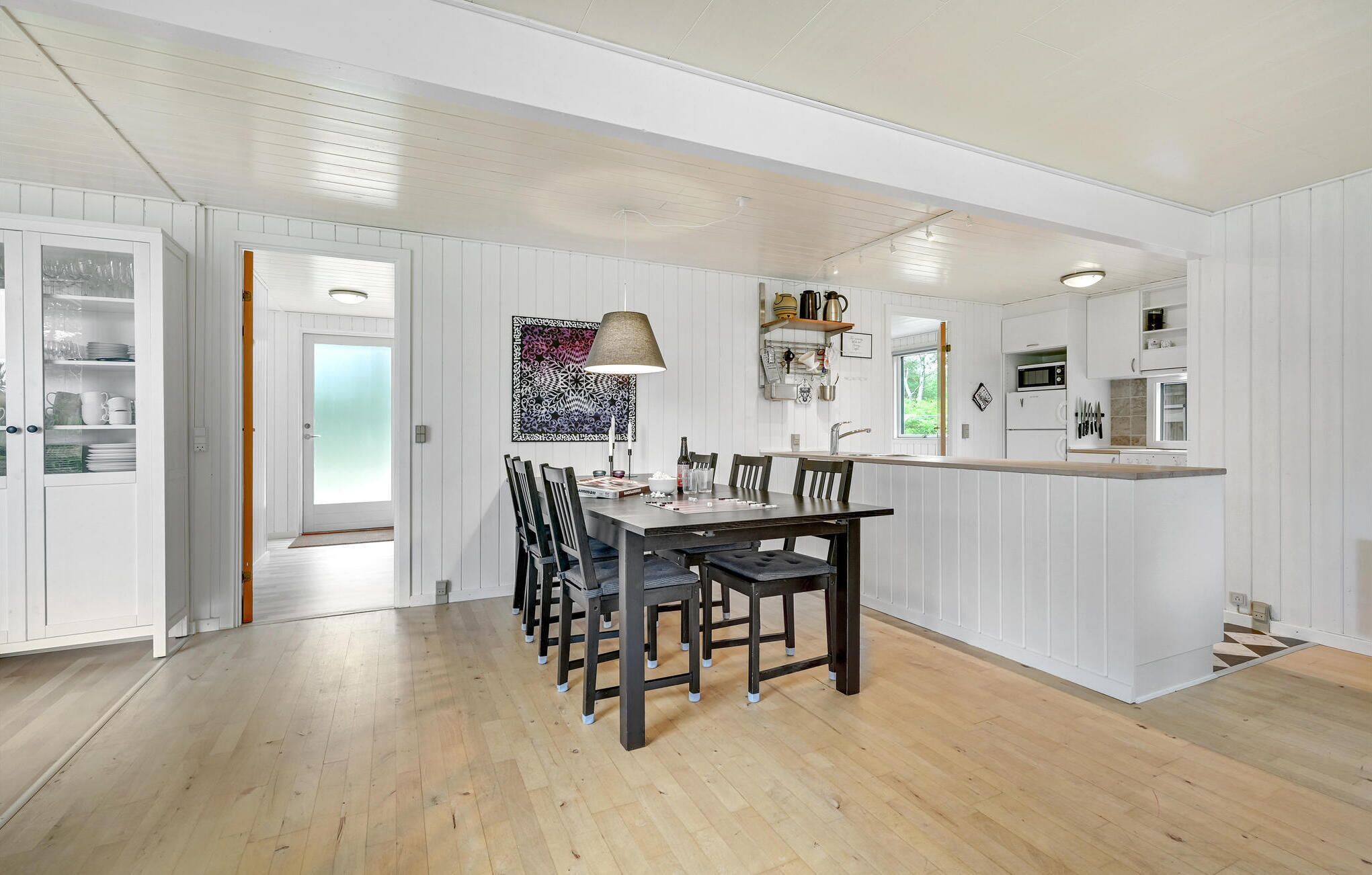 A modern kitchen with white cabinetry, a dining area with a wooden table and chairs, and a view of the outdoors through a door.