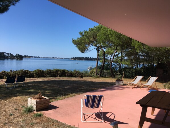 Eine Terrasse im Freien mit Blick auf einen See, zwei Liegestühle, ein Holztisch und eine kleine Holzhütte.